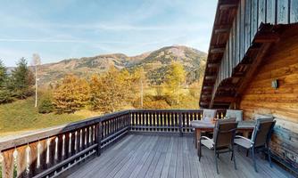 A cozy terrace with a wooden railing and chairs, surrounded by autumn scenery. In the background, mountains and trees with colorful foliage are visible.
