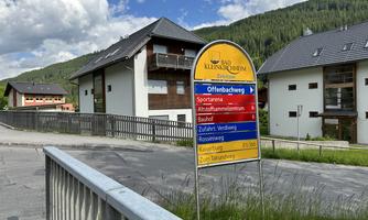 A directional sign in a quiet residential area with modern houses and green mountains in the background. The sky is partly cloudy.