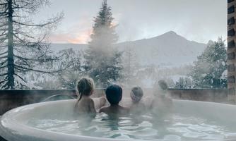 A group of people relaxes in a heated hot tub with a view of snow-covered mountains. The surroundings are romantic and filled with mist, creating a cozy atmosphere.