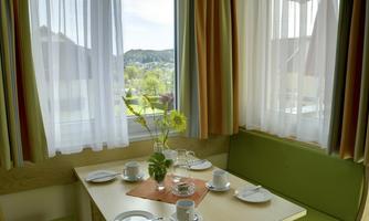 An inviting dining area with a table and set plates. Bright light comes through the windows, and you can see a green landscape outside.