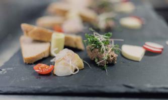 Ein ansprechend angerichteter Teller mit verschiedenen Häppchen, darunter Brot, Aufstrich und frisches Gemüse. Die Speisen sind auf einer schwarzen Platte präsentiert und wirken sehr appetitlich.
