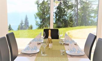 A festively set table with glasses and a bottle of wine. In the background, green trees and a beautiful view can be seen.