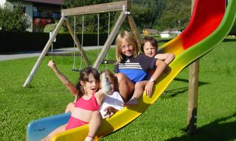 A joyful group of children is sliding down the slide together outdoors. In the background, a swing and many green meadows can be seen.