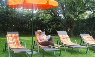 A woman is sitting relaxed in a lounge chair under an orange sun umbrella. In the background, other lounge chairs can be seen on a green lawn.