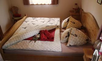A cozy bedroom with a wooden bed and colorful cushions. The blanket is thrown to the side, and fresh towels and a small bouquet of flowers are lying on the bed.
