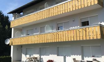 A modern building with two wooden balconies. The entrance area is surrounded by a well-maintained garden.