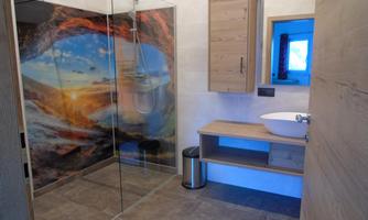 A modern bathroom with a glass shower and a sink on a wooden shelf. The wall behind the shower features a beautiful landscape painting with a sunset.