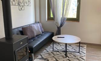 A cozy living room with a black sofa and a round table. The room has large windows with curtains and a fireplace in the corner.