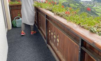A person is tending to plants on a balcony overlooking a green mountain landscape. It is a cloudy day, but the view is picturesque.