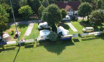 An aerial view of a campsite with motorhomes and tents in a green environment. In the background, a building and a sports field can be seen.