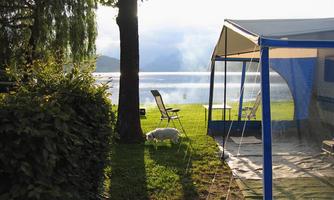 A picturesque campsite by the water with a view of a tranquil lake. In the background, there are chairs and green grass.
