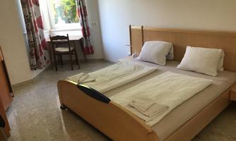 A simple bedroom with a double bed and two pillows. A table and a chair are situated next to a window with curtains.