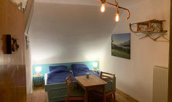 A cozy room with a double bed, two nightstands, and a dining table. A picture of a landscape hangs on the wall, and some wooden skis are decorated on the wall.