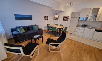 A modern living kitchen with a cozy sofa and a dining table. The decor is bright and friendly, featuring wooden flooring and elegant furniture.