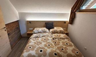 A cozy bedroom with a double bed and a floral bedspread. The room has large windows and modern wood paneling.