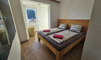 A simple bedroom with two single beds and wooden frames. The room has a window overlooking the outside and is bright and pleasantly furnished.