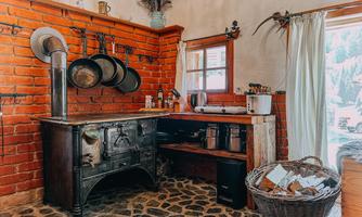 A rustic kitchen with red brick walls and a traditional stove. Pots hang on the wall and a basket stands next to the countertop.
