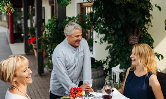 Two women are sitting at a beautifully set table outdoors, enjoying a meal. A waiter smiles and serves them wine.