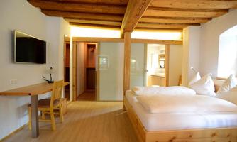 A cozy room with a wooden beam ceiling and modern furniture. There is a large bed, a table with a chair, and a TV on the wall.