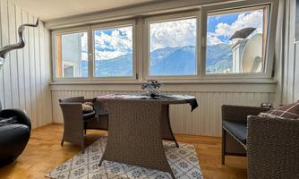 A cozy living room with a table and chairs made of rattan. Large windows offer a view of the mountains and the sky.