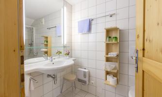 A modern bathroom with white tiles and elegant furnishings. There is a large mirror, sink, and a wooden shelf with toiletries.