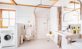 A bright bathroom with white tiles and wooden beams. It features a shower, a sink, and a washing machine.