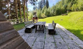A cozy seating area with a table and benches, surrounded by green grass and trees. In the background, there is a barbecue area.