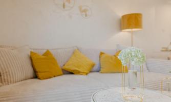 A cozy living room with a sofa and yellow cushions. A flower vase is on a table, and an elegant lamp shines in the background.