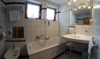 A modern bathroom with a bathtub and a sink. The walls are tiled with white tiles and there are natural light sources coming through the windows.