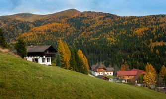 A picturesque mountain landscape with colorful autumn trees. In the foreground, traditional houses stand in a green meadow.