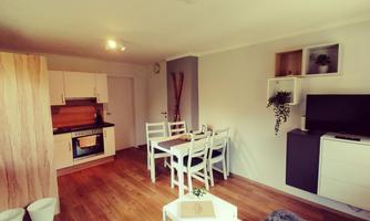 A modern living room with a bright kitchen and a dining table. The decor is simple and inviting, with wooden floors and cozy furniture.