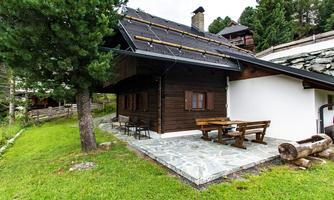 A cozy wooden house surrounded by green grass and trees. In front of the house is a terrace with a table and benches.