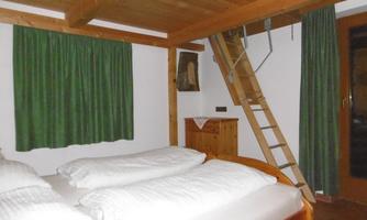 A cozy bedroom with two beds and wooden paneling. The windows are covered with green curtains, and a ladder leads to a sleeping loft.