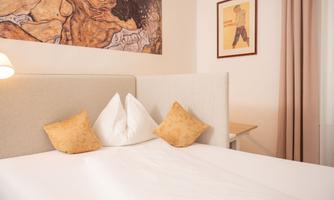 A cozy bedroom with a large bed and decorative pillows. An artwork hangs on the wall and there is a desk with a chair.