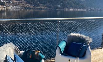 A cozy terrace with two chairs and a glass table. In the background, you can see a peaceful lake and mountains under a clear sky.