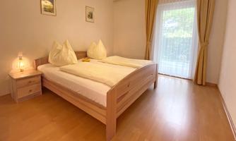 A cozy bedroom with a large bed and two pillows. The large windows let in a lot of light and provide a view outside.