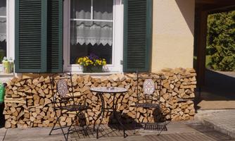 A cozy outdoor area with a small table and two chairs. In the background, stacked logs and a window with green shutters can be seen.