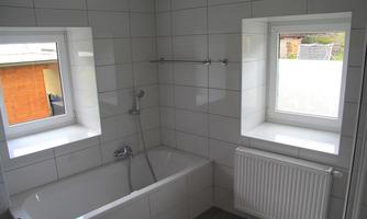 A modern bathroom with a bathtub and two windows. The walls are tiled and the rooms feel bright and welcoming.