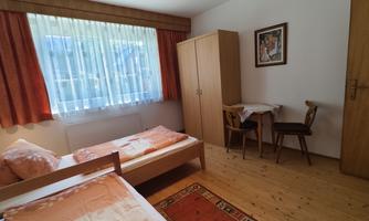 A simple bedroom with two beds, a table and chairs. Large windows let in a lot of light.