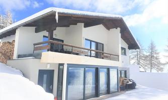 A beautiful modern chalet in the snow. It has large windows and a balcony overlooking the winter landscape.