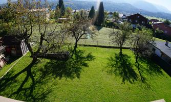 A green meadow with several trees and long shadows. In the background, gentle hills and houses can be seen.