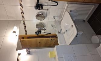 The image shows a modern bathroom with white tiles and a sink. In the background, a mirror and a wooden double door can be seen.