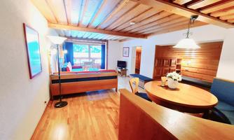 A cozy living space with a wooden ceiling and warm wooden floors. The room features a table, chairs, and a relaxed seating area with a view of a window.