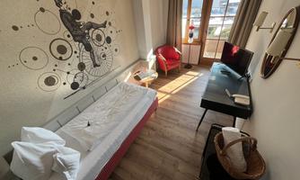 A modern furnished room with a bed, a desk, and a red armchair. The walls are decorated with a creative wall motif and there is plenty of daylight.