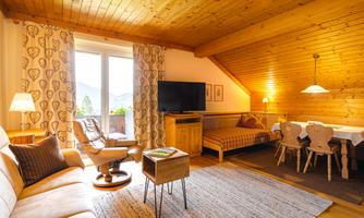 A cozy living room with wooden walls and a large window. There is a comfortable seating area, a table, and a television.