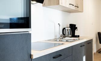 A modern kitchen with black and wood-colored cabinets. The room is bright and well-equipped with a sink, a coffee machine, and a refrigerator.