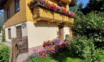 A beautiful wooden cottage with blooming flowers and an inviting garden. The balcony is abundantly decorated with plants, creating a friendly atmosphere.