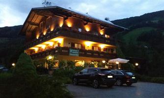 A charming, illuminated alpine-style hotel surrounded by green hills. Several cars are parked in front of the hotel.