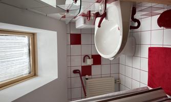 A small bathroom with white and red tiles. There is a window, a shower, and basic sanitary facilities.