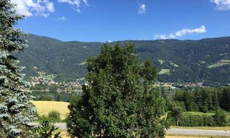 A picturesque landscape with mountains in the background and a clear blue sky. In the foreground, green trees and fields stand.